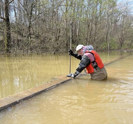 A water expert wades in floodwaters and uses equipment that looks like a tall metal pole to measure the height of floodwaters