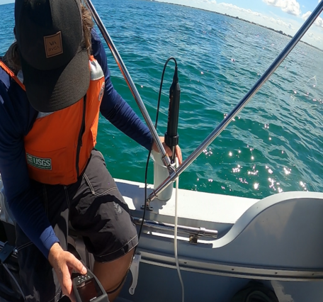 a scientist sits on the edge of a boat on the ocean with an instrument submerged in a sample tube of water
