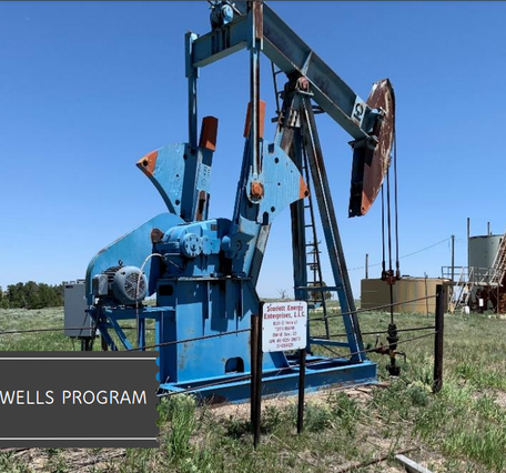 Image shows a rusting oil & gas pumpjack on a grassy field with a petroleum storage facility with a blue sky