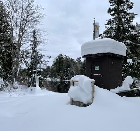 A brown streamgage housing with two feet of fresh snow piled on the roof with a cableway in the background