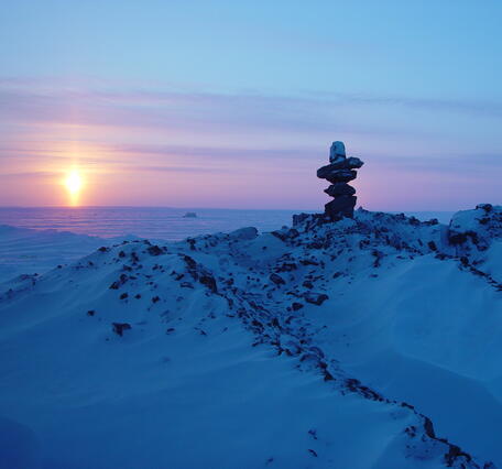 Image shows a rock cairn on a snowy peak with the sun on the horizon